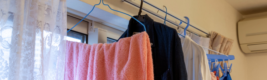 窓際のカーテンレール付近にハンガーを掛けた室内干しの様子。専用の物干しスペースがない場合の、日常的な洗濯物の悩みや課題を表現。